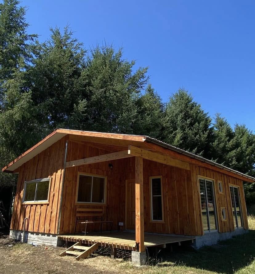 Cabaña de madera con porche, banca y acceso cubierto entre los árboles