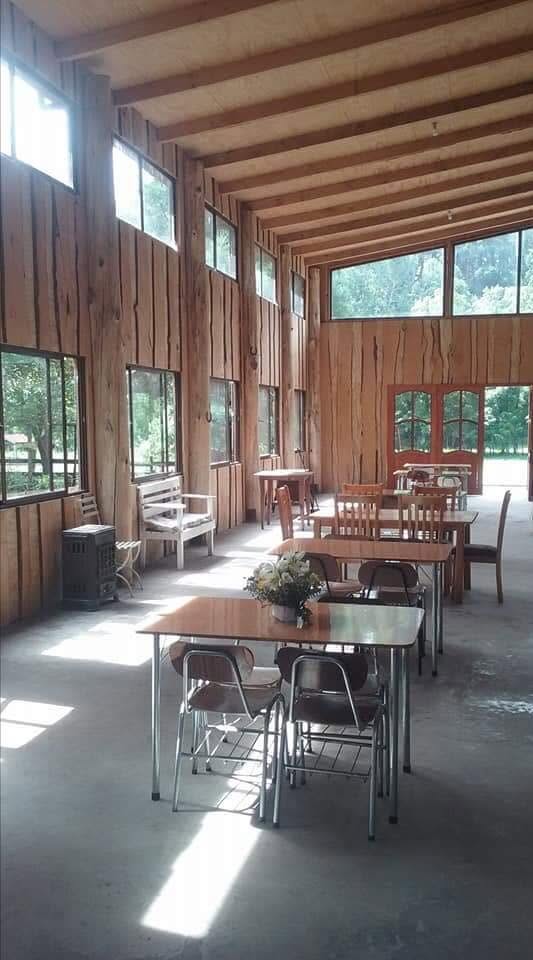 Salón campestre con paredes de madera, grandes ventanas y mesas para encuentros